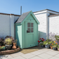 Green garden shed with a black roof against a white brick wall.